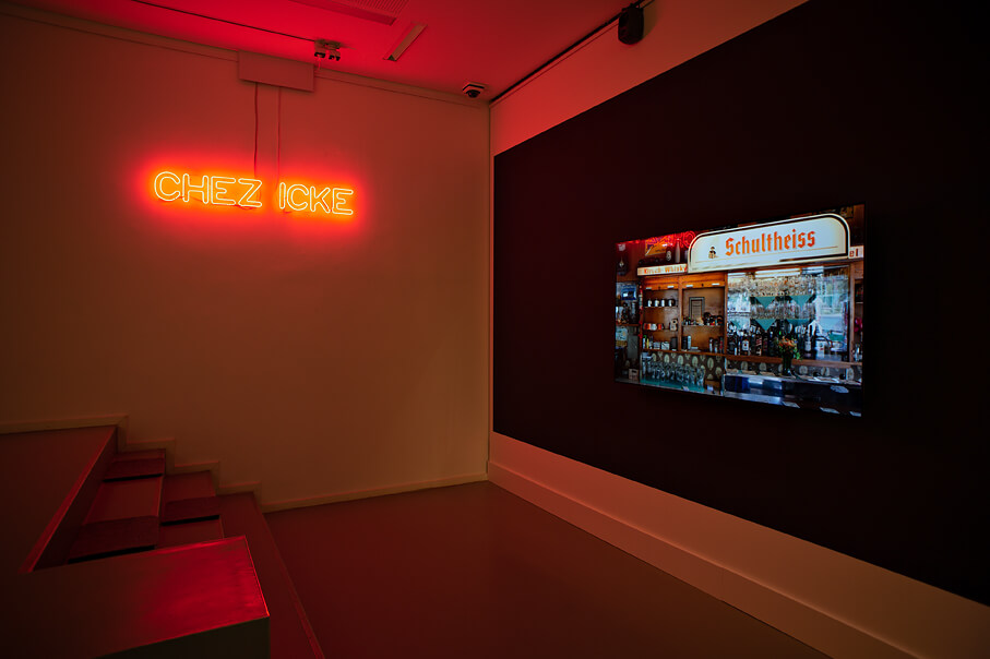Fotografie eines LED-Neon Schriftzuges des Ausstellungstitels: "Chez Icke" am linken Bildrand, sowie eine über den Beamer abgespielte Kneipenszene am rechten Bildrand 